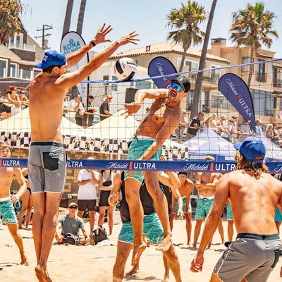 Beach volleyball game with players on a sandy court, palm trees, and buildings in the background.