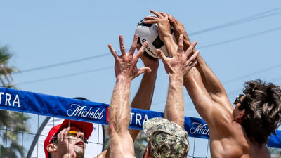 Three men playing beach volleyball reaching up to hit the ball over a net with a clear blue sky in the background.
