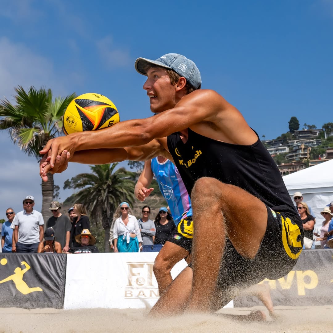 Beach volleyball player in action on a sandy court with spectators and palm trees in the background.